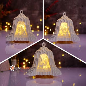 May include: Three white paper Christmas bell-shaped lanterns with intricate cut-out designs. Each lantern has a warm, glowing light inside, showcasing different holiday scenes: a Christmas tree, a snowman, and deer in a forest. The lanterns sit on a brown base.