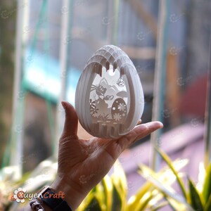 May include: A white papercraft Easter egg with a 3D design featuring a bunny rabbit, flowers, and a mountain scene. The egg is intricately cut and layered, creating a delicate and intricate design.