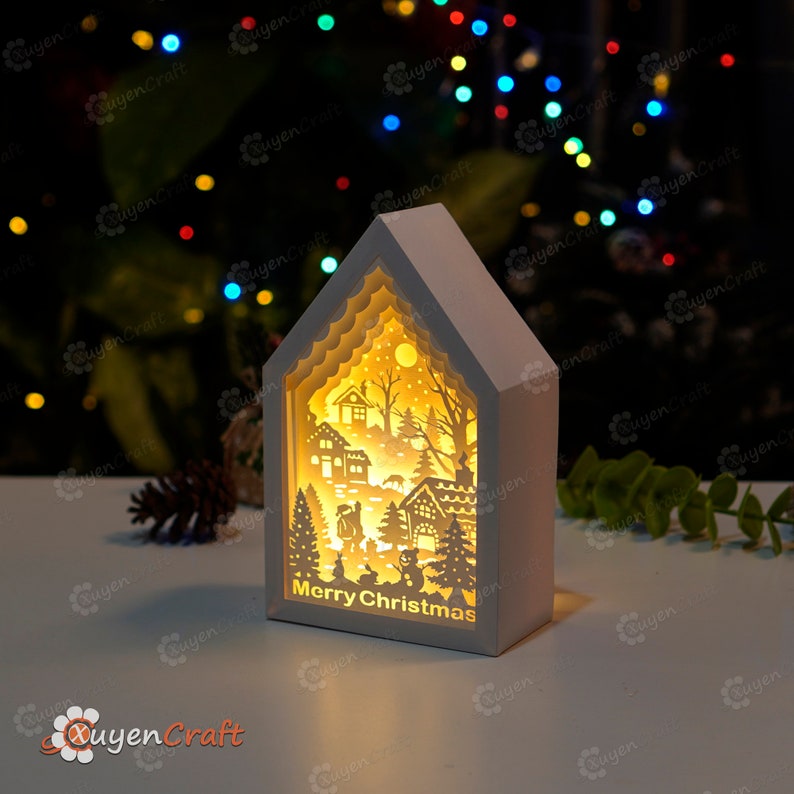 May include: A white wooden house-shaped light-up ornament with a cut-out winter scene and the words "Merry Christmas". The scene includes a snowman, a deer, a house, and a Christmas tree.