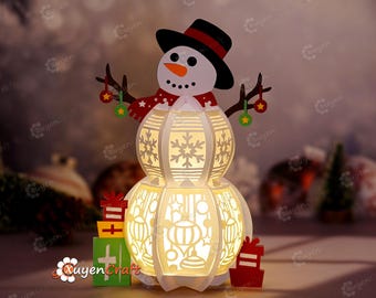 May include: A white papercraft snowman with a red and white scarf, black hat, and a light inside. The snowman is holding two green and red ornaments. The snowman is sitting on a table with two red and green wrapped gifts.