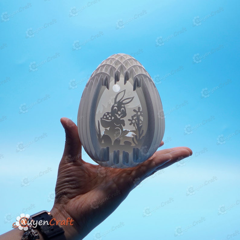 May include: A white papercraft Easter egg with a cutout design of a bunny holding a basket of eggs. The egg is held in a person's hand against a light blue background.