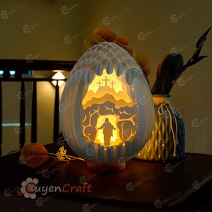 May include: A white egg-shaped paper lamp with a cut-out design depicting a scene from the Bible. The scene shows a figure emerging from a cave with a cross behind them. The lamp is lit from within, casting a warm glow.