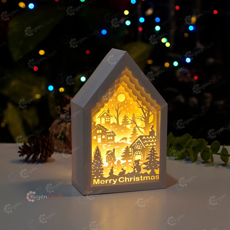 May include: A white wooden Christmas house with a cut-out design of a winter scene. The house is illuminated from within, creating a warm glow. The text "Merry Christmas" is visible at the bottom of the house.