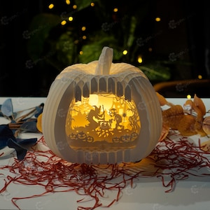 May include: A white papercraft pumpkin with a light inside, depicting a Halloween scene with a witch, a cauldron, and a cat. The pumpkin is sitting on a bed of red and white shredded paper.