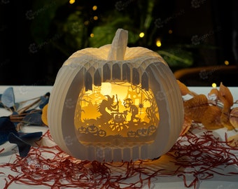 Może przedstawiać: Biała papierowa dynia z wbudowanym światłem, przedstawiająca scenę z Halloween z czarownicą, kotłem i kotem. Dynia stoi na łóżku z czerwono-białego posiekanego papieru.
