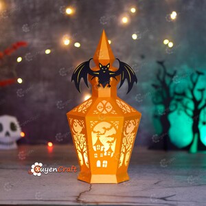 May include: An orange paper lantern with a bat silhouette on top, designed for Halloween. The lantern features cut-out designs of trees, a haunted house, and bats. The lantern is lit from within, creating a warm glow.