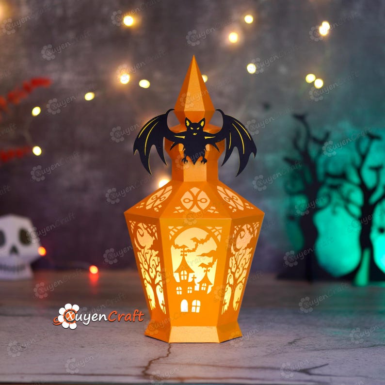 May include: An orange paper lantern with a bat silhouette on top, designed for Halloween. The lantern features cut-out designs of trees, a haunted house, and bats. The lantern is lit from within, creating a warm glow.
