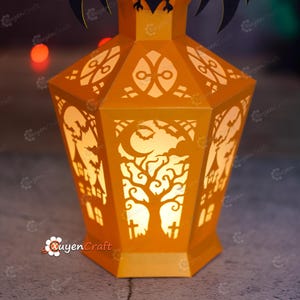 May include: An orange paper lantern with intricate cut-out designs for Halloween. The lantern features a tree, moon, bats, and haunted house silhouettes. The lantern is lit from within, creating a warm glow.