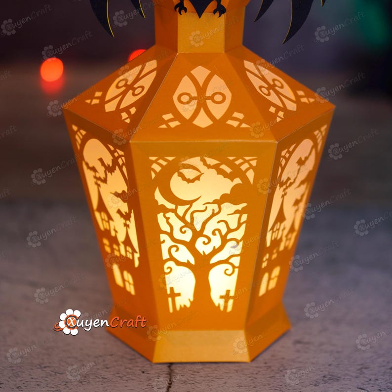 May include: An orange paper lantern with intricate cut-out designs for Halloween. The lantern features a tree, moon, bats, and haunted house silhouettes. The lantern is lit from within, creating a warm glow.