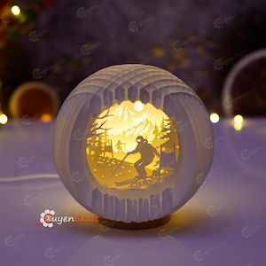 May include: A spherical white paper-cut lamp featuring an illuminated interior scene of a skier on a mountain slope. The layered design creates a 3D effect with silhouettes of trees and a skier against a warm yellow light, set on a circular base.