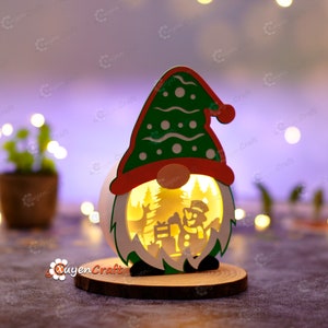 Pode incluir: Enfeite de Natal em madeira em forma de gnomo, com chap&eacute;u verde e borda vermelha. O corpo branco do gnomo apresenta um desenho recortado que mostra um boneco de neve, veados e &aacute;rvores, iluminados por dentro. Apoia-se numa base de madeira.