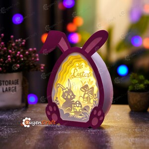 May include: A pink and white 3D papercraft Easter bunny lamp with a light inside. The bunny is holding an egg and there is a sign that says "Easter" inside the egg.