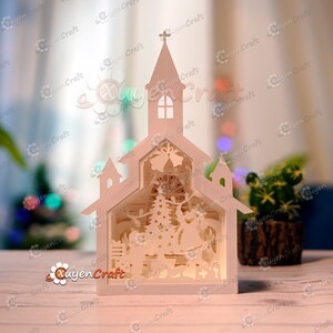 May include: White papercraft model of a church with a Christmas scene inside. The church has a steeple with a cross on top and a window. The scene inside the church includes a Christmas tree, a Santa Claus figure, and other Christmas decorations.