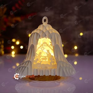 May include: A white paper Christmas bell-shaped lantern with a cutout nativity scene illuminated from within. The lantern has a textured surface and a wooden base. The background is blurred with warm, golden lights.