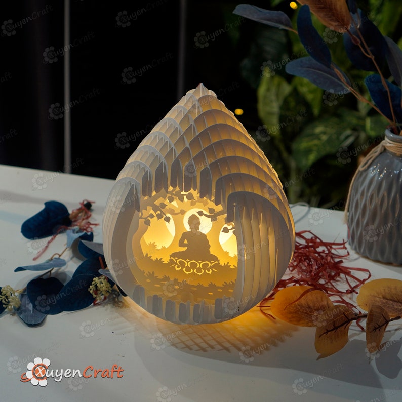 Op de afbeelding: Een witte papieren lamp in de vorm van een traan met een uitgesneden ontwerp van een mediterend Boeddhabeeldje erin. De lamp is aan en geeft een warm licht.