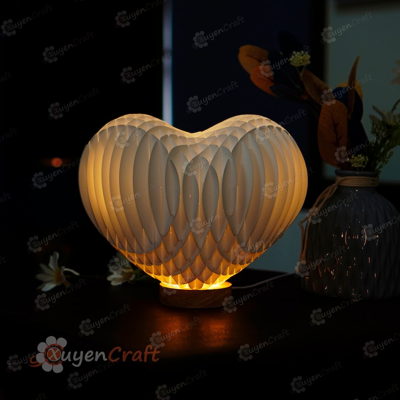 May include: A white heart-shaped paper lamp with a warm glow. The lamp is on a wooden base and is lit up, casting a soft light.
