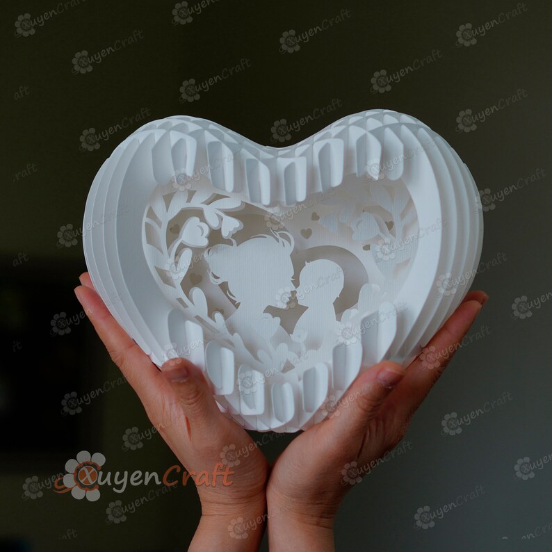 May include: White papercraft heart-shaped sculpture with a cutout design of a mother and child. The sculpture is held in two hands.