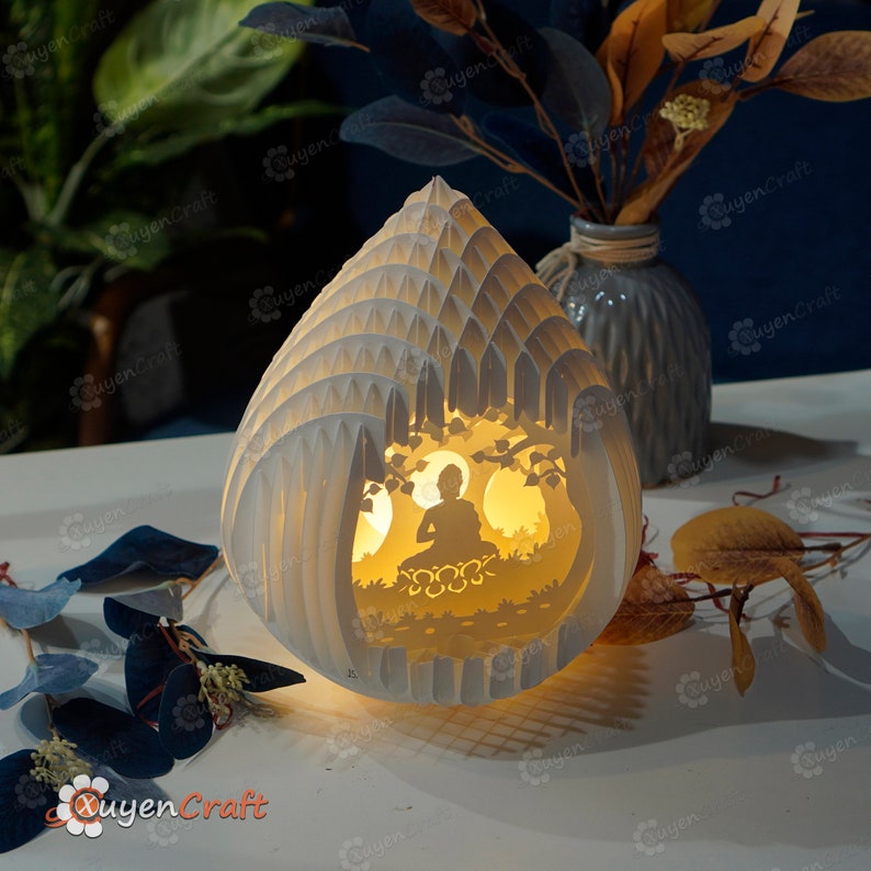 Op de afbeelding: Een witte papieren lamp in de vorm van een traan met een uitgesneden ontwerp van een mediterend Boeddha-figuur erin. De lamp wordt van binnenuit verlicht en geeft een warm gloed.