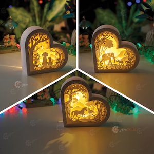 May include: Three white heart-shaped wooden night lights with intricate laser cut designs. Each light features a different scene: a dog and cat in a forest, an elephant and a baby elephant in a jungle, and a horse and foal in a forest. The lights are illuminated from within, casting a warm glow.
