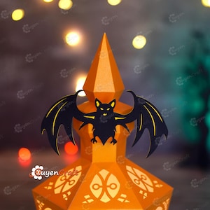 May include: A Halloween-themed paper lantern in orange with a black bat silhouette on top. The lantern has intricate cut-out designs and a pointed top. The background features blurred lights, creating a festive atmosphere.