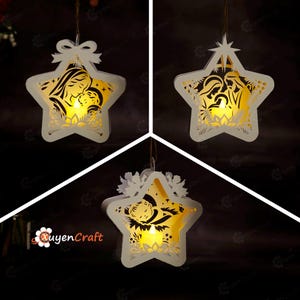 May include: Three white star-shaped hanging ornaments featuring intricate laser-cut nativity scenes illuminated by warm yellow internal lights. Each ornament displays a different religious figure design and hangs from a thin brown string.