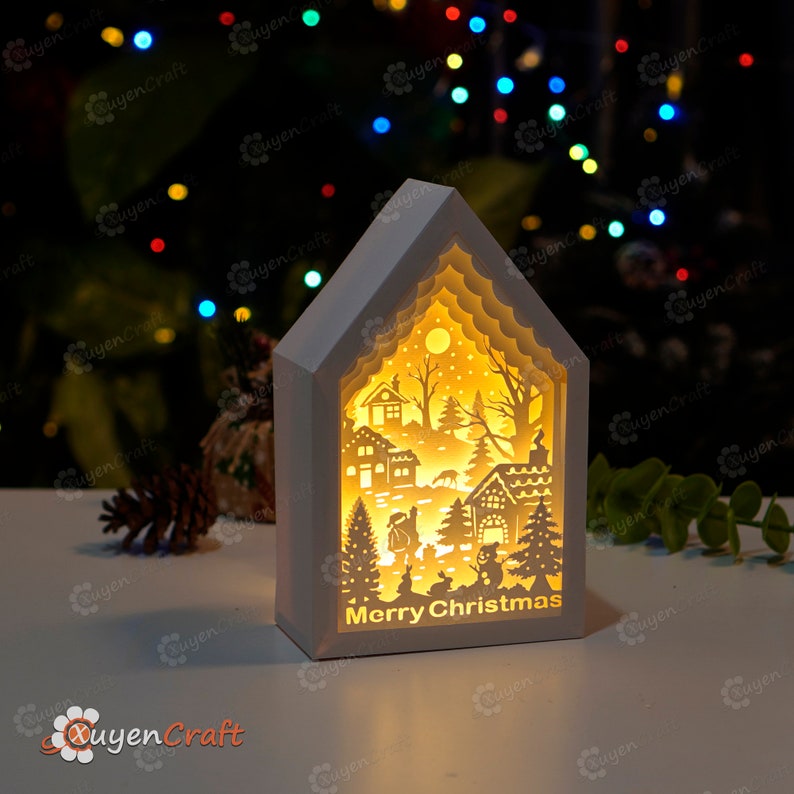 May include: A white wooden Christmas village scene with a light inside. The scene features a snowy village with houses, trees, and a deer. The text "Merry Christmas" is printed on the bottom of the scene.