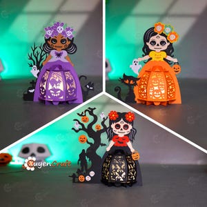 May include: Three paper cut-out Halloween decorations. Each features a stylized figure with a floral crown and a lantern-like skirt. The lanterns have cut-out designs of bats, pumpkins, and other Halloween motifs. The figures are in purple, orange, and black.