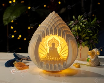 May include: A white papercraft lamp shaped like a lotus flower with a silhouette of a meditating Buddha inside. The lamp is lit and casting a warm glow.