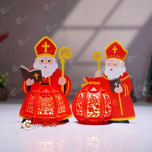 May include: Two red paper cut-out figures of St. Nicholas holding a staff and a book with a cross. One figure is next to a red lantern with a Christmas tree design. The figures wear red robes and hats with a gold cross.