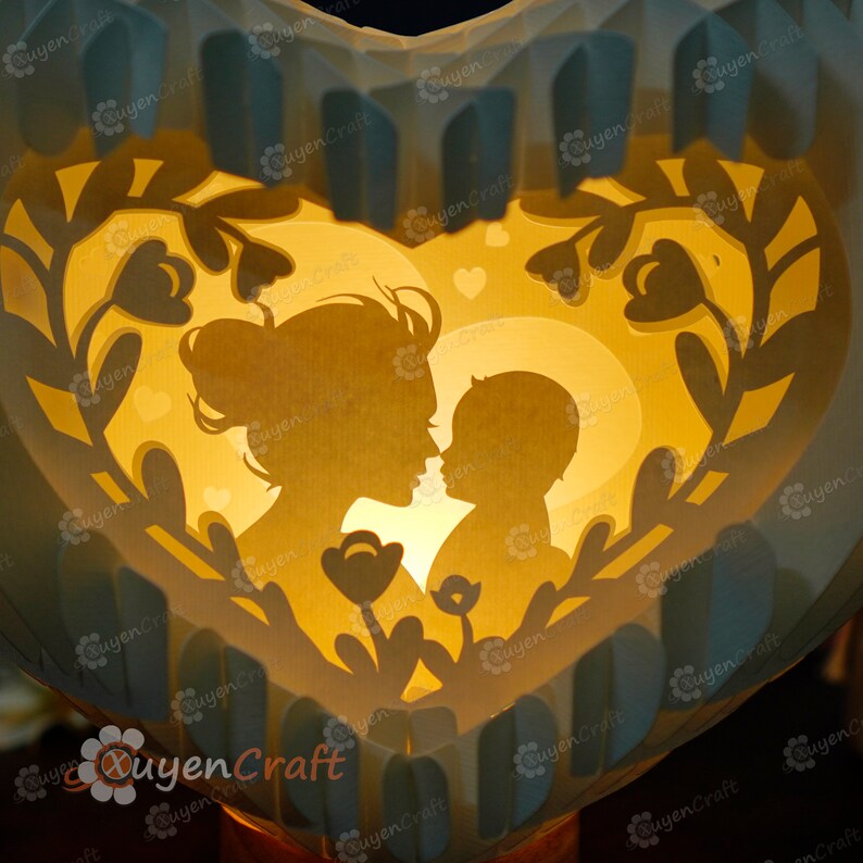 May include: A white heart-shaped paper lamp with a cutout design of a mother and child. The lamp is illuminated from behind, creating a warm glow. The design features floral elements and small hearts.