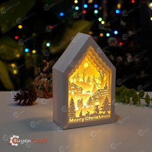 May include: A white wooden house-shaped light-up Christmas decoration with a cut-out design of a winter village scene. The light illuminates the scene, creating a warm glow. The words "Merry Christmas" are printed on the bottom of the house.