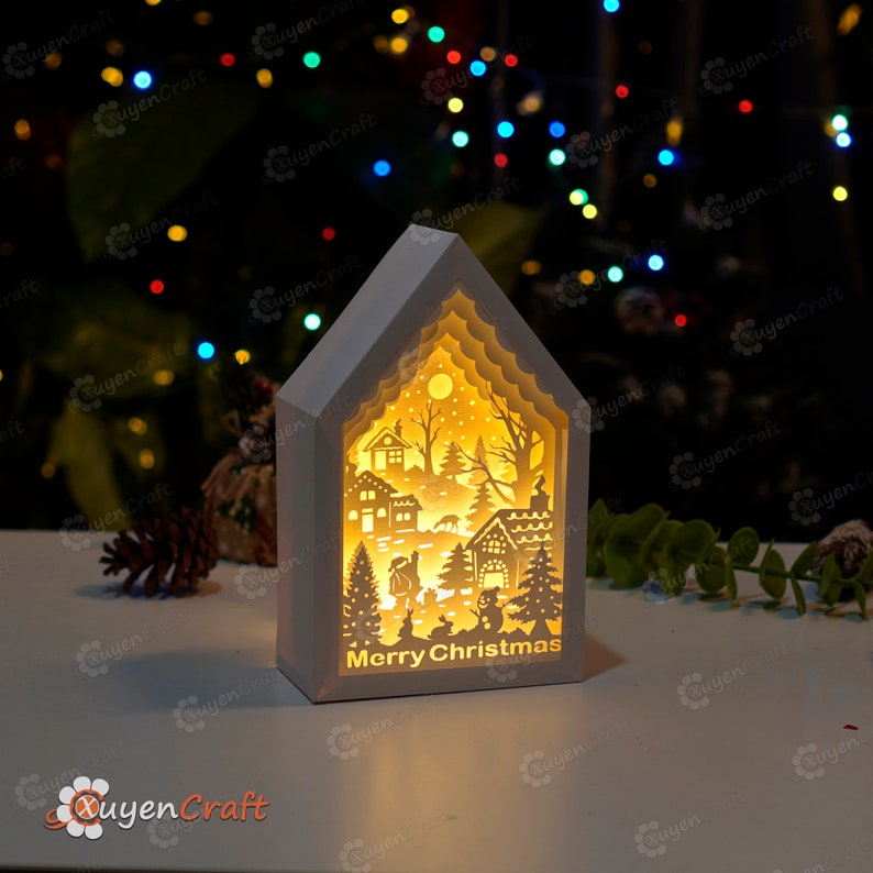 May include: A white wooden house-shaped light-up ornament with a cut-out design of a winter village scene. The ornament has a warm yellow glow and the words "Merry Christmas" are printed on the bottom.