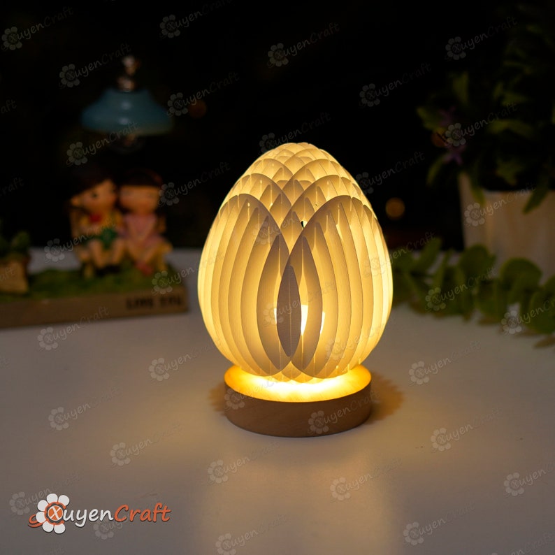 May include: A white, wooden, egg-shaped lamp with a warm, yellow light emanating from the base. The lamp is made of thin, overlapping pieces of wood, creating a delicate and intricate design.