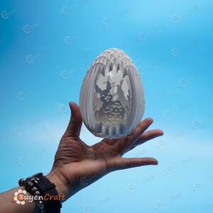 May include: A white papercraft Easter egg with a 3D design of bunnies and an Easter basket. The egg is held in a person's hand against a blue background.