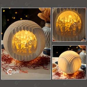 May include: A white paper lamp shaped like a sphere with a cut-out design of a winter village scene. The lamp is lit and casts a warm glow on the scene. The lamp is sitting on a table with red and brown paper shreds.