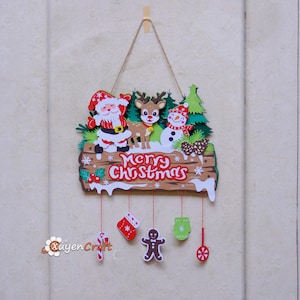 May include: A decorative wooden Christmas wall hanging featuring Santa Claus, a reindeer, and a snowman above a sign that reads Merry Christmas. Five small holiday-themed ornaments, including a candy cane, stocking, gingerbread man, mitten, and peppermint, hang from the base by thin red strings.