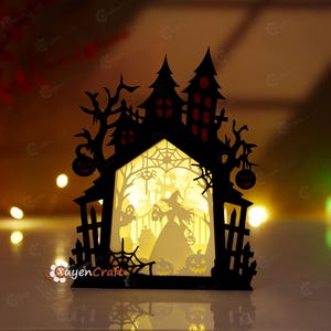 Puede incluir: Una caja de luz de papel recortado negro con la silueta de una casa encantada, una bruja, un fantasma y calabazas. El interior está iluminado con un cálido resplandor amarillo, resaltando los intrincados recortes de telarañas y ramas.