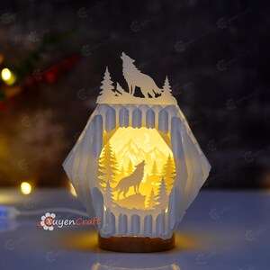 Może przedstawiać: Biały lampion z wycinanego papieru z warstwowym wzorem przedstawiającym sylwetkę wyjącego wilka na górze oraz podświetloną scenę wewnątrz z wilkiem, sosnami i górami. Konstrukcja ma sześciokątny kształt z misternymi wycięciami i ciepłym światłem.