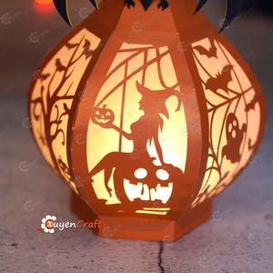 May include: Orange paper lantern with Halloween-themed cutouts, including a witch, bats, and a jack-o'-lantern. The lantern is lit from within, creating a warm glow. The text "Quyen Craft" is visible.