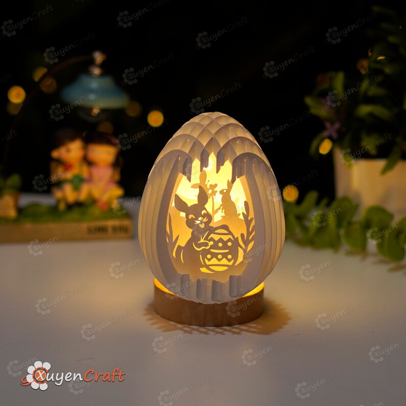 May include: A white paper Easter egg shaped night light with a cut-out design of a bunny holding an Easter egg. The night light is illuminated with a warm white light.