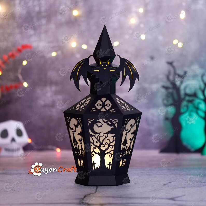 May include: Black Halloween lantern with a bat and tree design. The lantern has a pointed top and intricate cutouts of a tree, houses, and a moon. The lantern is lit from within, creating a spooky glow. The lantern is made of paper.