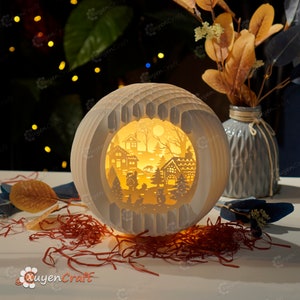 May include: A white papercraft lamp with a winter village scene inside. The lamp is lit and the scene is illuminated with a warm glow. The lamp is sitting on a table with red shredded paper.
