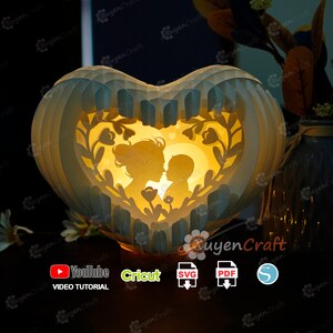 May include: A heart-shaped papercraft lamp with a mother and child silhouette inside. The lamp is lit from within, casting a warm glow. The lamp is decorated with floral designs and has a white and blue color scheme. The image includes the text "YouTube Video Tutorial", "Cricut", "SVG", "PDF", and "S".