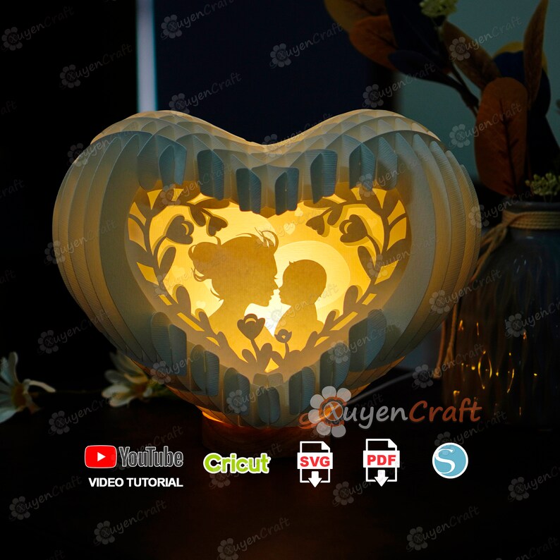 May include: A heart-shaped papercraft lamp with a mother and child silhouette inside. The lamp is lit from within, casting a warm glow. The lamp is decorated with floral designs and has a white and blue color scheme. The image includes the text "YouTube Video Tutorial", "Cricut", "SVG", "PDF", and "S".