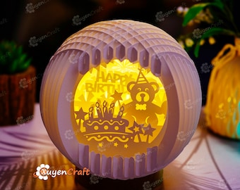 May include: A white paper sphere with a cut-out design of a birthday cake, candles, stars, and a teddy bear wearing a party hat. The sphere is illuminated from within, casting a warm glow. The text "Happy Birthday" is visible in the cut-out design.