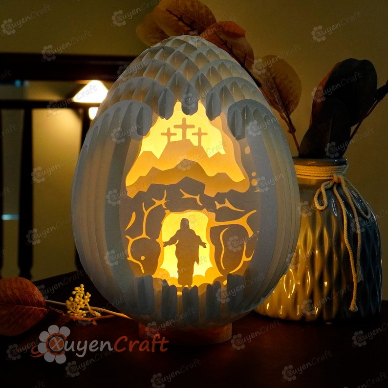 May include: A white ceramic egg-shaped lamp with a cutout design depicting a scene of Jesus emerging from a tomb. The lamp is illuminated from within, casting a warm glow. The scene includes a cross and a mountain.