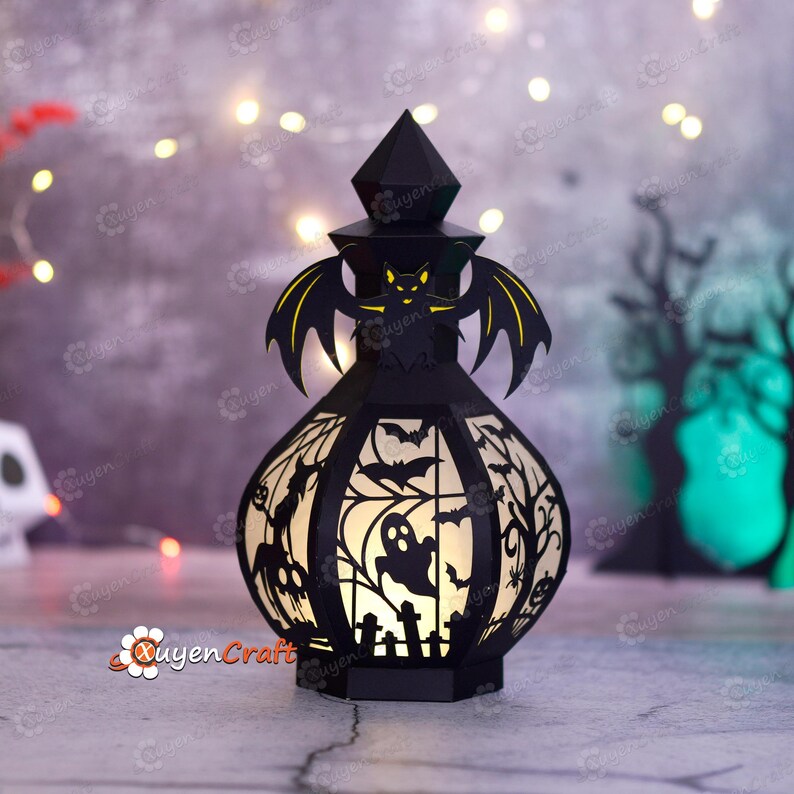 May include: Black Halloween lantern with a bat and spooky silhouettes. The lantern features a geometric design with cut-out bats, ghosts, and trees. The lantern is lit from within, creating a warm glow. The lantern is a decorative item for Halloween.