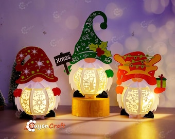 May include: Three Christmas gnomes with light-up features. The gnomes are made of paper and have a white, paper lantern inside. The gnomes are wearing red, green, and yellow hats. The gnome on the left is holding a sign that says "Xmas".
