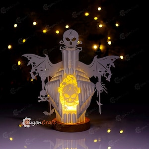 May include: A white, layered Halloween-themed lantern with a skull and crossbones design. The lantern features bat wings, a spider, and a skull with an X on its forehead. The lantern is illuminated from within, creating a warm glow. The base is made of wood.