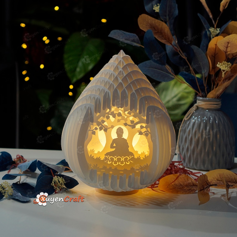 Op de afbeelding: Een witte lamp van papierkunst in de vorm van een traan met een uitsnijding van een mediterend Boeddhabeeldje erin. De lamp wordt van binnenuit verlicht en geeft een warm gloed.